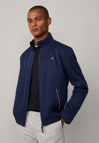 Chaqueta ligera de color azul marino con un cuello alto, cremallera frontal y dos bolsillos laterales con acentos beige. Fabricada con tejido suave.