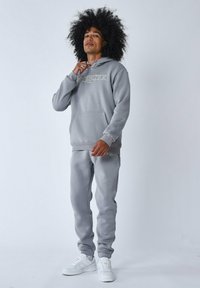 Sudadera gris y pantalones de chándal a juego, hechos de material suave, con un logo en el pecho. Llevados con zapatillas blancas, sobre un fondo liso.