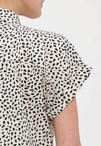 Chemise à manches courtes, avec col, en tissu léger, avec un fond crème et un motif de taches abstraites noires éparpillées.