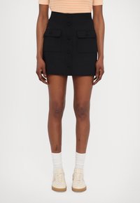 Zwarte mini-jurk met een hoge taille, voorzien van een knoopsluiting aan de voorkant, twee grote klepzakken en een gladde textuur. Gecombineerd met lichte sneakers.