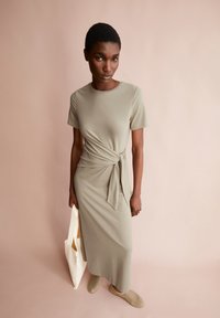 Salbeigrünes, kurzärmliges Kleid in knieumspielender Länge mit einem gebundenen Taillendetail, aus glattem Stoff gefertigt. Kombiniert mit hellen Slipper-Schuhen und einer Tragetasche.