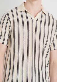 Beżowa polo z krótkim rękawem, z pionowymi czarnymi paskami i teksturowanym wzorem. Posiada spiczasty kołnierzyk i dekolt w kształcie litery V.