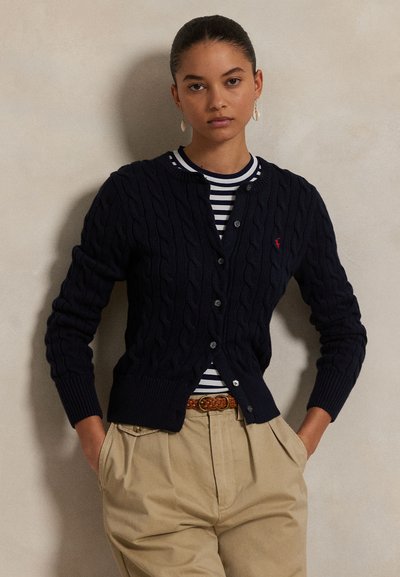 Cardigan en maille torsadée bleu marine avec boutons sur toute la partie avant, poignets et ourlet côtelés ; porté sur un T-shirt rayé, associé à un pantalon beige et une ceinture tressée.