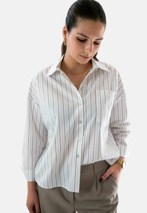 Chemise blanche à manches longues avec des rayures verticales en brun clair, dotée d'une poche et d'un col classique, associée à un pantalon beige.