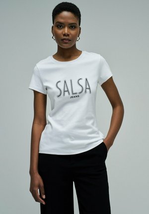 Mujer con cabello corto, llevando pendientes de aro dorados, una camiseta blanca ajustada con el texto "SALSA JEANS" y pantalones negros, posando con una mano en el bolsillo.