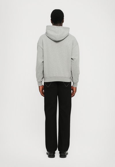 Haut à capuche en coton gris avec une couture au dos, une capuche spacieuse et des poignets côtelés, associé à un pantalon noir à jambe droite et des chaussures noires.