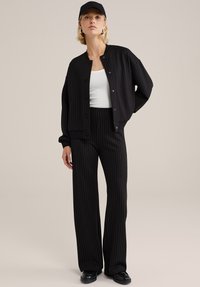 Črno bomber jakna s tanjšimi črtami čez belo telo, kombinirana z ujemajočimi širokimi hlačami in črnimi loafer čevlji, na vrhu pa črna kapa.