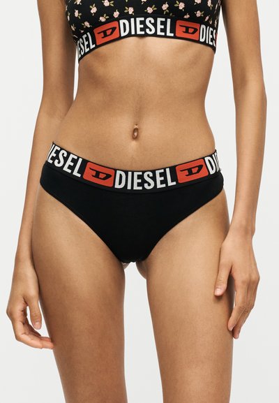 Femme portant un crop top noir à fleurs et un sous-vêtement noir avec de larges bandes élastiques marquées "DIESEL" en lettres blanches et rouges.
