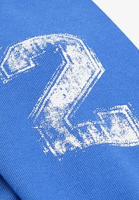 Tessuto di cotone blu con una grande stampa del numero "2" bianco, testurizzato e con un effetto consumato, dalla texture liscia e a maglia.