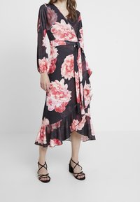Blommig omslagsklänning med långa ärmar, svart bakgrund, stort rosa piontryck, volangkant, knyts i midjan, kombinerad med svarta sandaler med remmar.