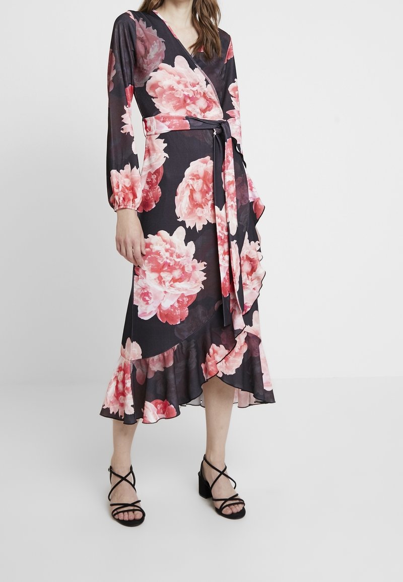 Blommig omslagsklänning med långa ärmar, svart bakgrund, stort rosa piontryck, volangkant, knyts i midjan, kombinerad med svarta sandaler med remmar.