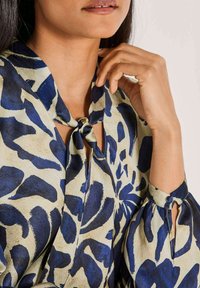 Frau mit einer beigen Bluse mit großen blauen abstrakten Blumenmustern, einem Neckholder-Detail und geknöpften Puffärmeln.