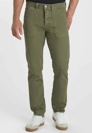 Man draagt olijfgroene broek met voorzakken en witte sneakers, staand tegen een effen achtergrond.