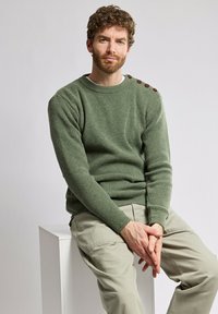 Zielony sweter dzianinowy o rypsowej fakturze i okrągłym dekolcie, z brązowymi guzikami na lewym ramieniu; zestawiony z jasnobeżowymi spodniami.