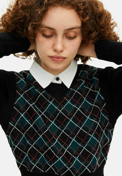 Jovem mulher com cabelo encaracolado e vermelho, vestindo um colete preto com padrão argyle em verde, branco e vermelho, por cima de uma camisa branca, com as mãos atrás da cabeça.