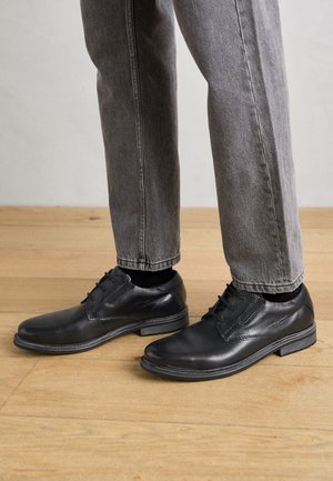 Schwarze, glatte Lederschuhe mit Schnürsenkeln und runder Spitze, kombiniert mit grauen Denim-Hosen und schwarzen Socken.