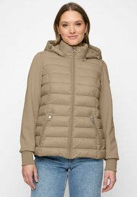 Frau in einer beigefarbenen, gesteppten Jacke mit Kapuze und Reißverschlusstaschen, kombiniert mit hellblauen Jeans, steht vor einem weißen Hintergrund.