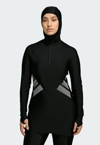 Sort atletisk tunika med høj hals og lynlås, med teksturerede grå diagonale detaljer på siderne. Lange ærmer med hul til tommelfingrene.