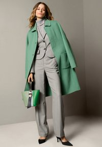 Groene wollen jas, grijs gestippeld pak, zwarte hakken, met een kleurgebroken handtas in groen en zwart. Gladde texturen, aansluitend ontwerp.