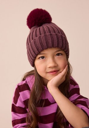 Niña joven con un gorro de punto morado con pompón y un suéter a rayas a juego, apoyando su rostro en la mano con una ligera sonrisa.
