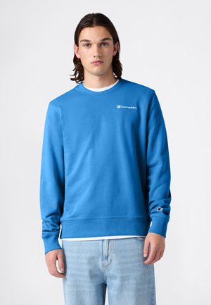 Junger Mann mit schulterlangem braunem Haar, der einen blauen Champion-Sweatshirt und hellblaue Jeans vor einem weißen Hintergrund trägt.