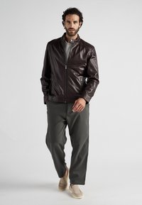 Brun läderjacka med dragkedja framtill och hög krage, tillsammans med olivgröna byxor och beigea slip-on skor; slät textur.