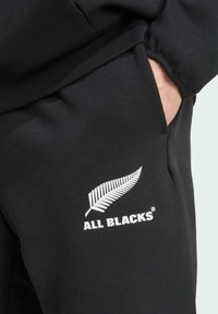 Crne trenirke od mekanog materijala s bijelim logotipom paprati i tekstom "ALL BLACKS" na lijevom džepu.