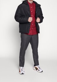 Man in een zwarte pufferjack over een rood overhemd, grijs geruite broek en witte sneakers met zwarte en gele details.