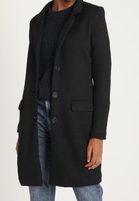 Cappotto nero textured con rever, due tasche frontali e chiusura a tre bottoni, indossato sopra un maglione scuro e jeans blu.