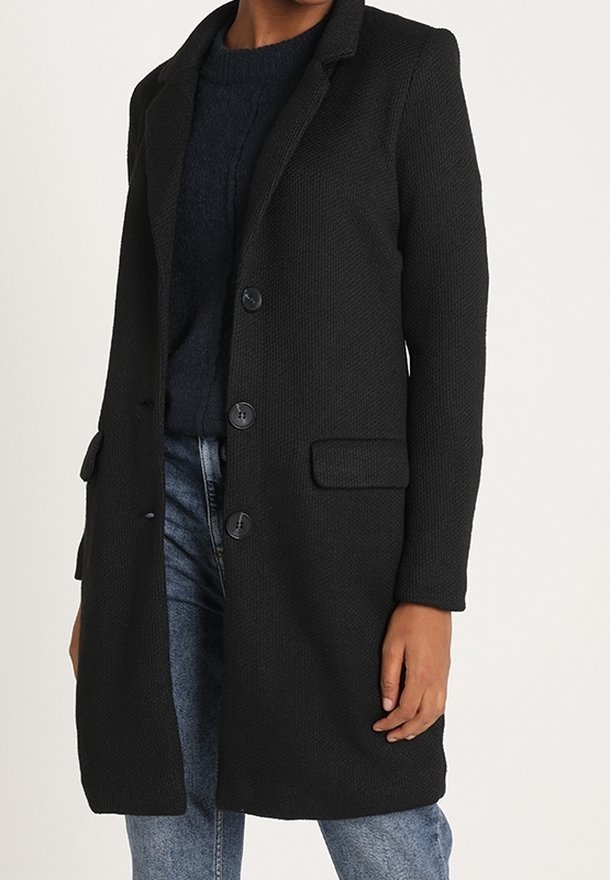 Cappotto nero textured con rever, due tasche frontali e chiusura a tre bottoni, indossato sopra un maglione scuro e jeans blu.