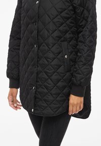 Kvalitna črna jakna s prešito površino, z gumbi na pritisnjenje, stranskimi žepi in rebrastimi manšetami. Ima gladko, sijajno površino in zaobljen rob.