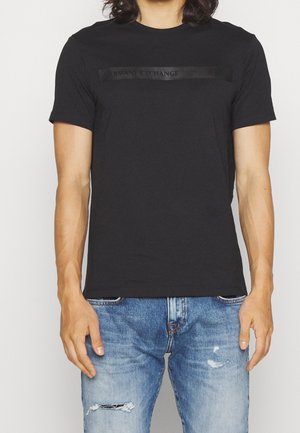 Schwarzes Baumwoll-T-Shirt mit kurzen Ärmeln und Rundhalsausschnitt. Mit einem strukturierten schwarzen Logo-Band über der Brust. Kombiniert mit abgenutzten blauen Jeans.