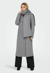 Cappotto grigio in lana con design aperto e ampio colletto scialle. Abbinato a una sciarpa coordinata e pantaloni neri a gamba larga. Texture liscia.