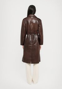 Femme aux longs cheveux bruns, portant un manteau en cuir marron foncé ceinturé et un pantalon large blanc, se tenant de dos contre un fond uni.