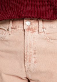 Pantalon en velours côtelé rose doux, avec fermeture à boutons, poche avant, et texture côtelée verticale. Porté avec un pull en maille bordeaux.