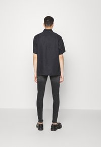 Camisa preta de mangas curtas feita de um tecido leve, combinada com jeans escuros de corte slim e sapatos pretos, vista por trás.