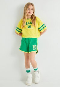 Keltainen lyhyt t-paita, jossa on vihreät raidat ja teksti "BRAZIL"; vihreät shortsit, joissa on valkoinen reunus ja "10" printti; valkoiset korkeat tennarit.