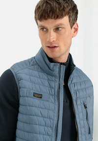 Blauer gesteppter Weste mit Stehkragen, Reißverschluss und einem strukturierten marineblauen Pullover darunter. Mit einem Logopatch und einer Seitentasche.