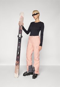 Ski gear featuring a black long-sleeve top, light pink snow pants with grey accents, and black ski boots. A patterned ski is held in one hand.