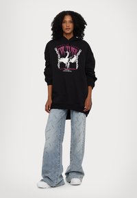 Black hoodie with graphic print featuring a swan and text, paired with light blue wide-leg jeans with repeated logo pattern. White sneakers.