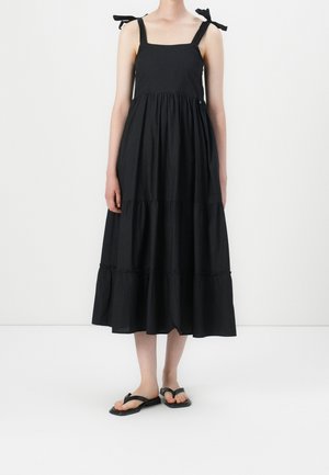 Černé midi šaty bez rukávů s vrstvenou sukní a zavazovacími ramínky, nošené s černými nízkými sandály, zobrazené na modelce na bílém pozadí.