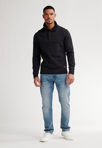 Man staat in een zwart knooptrui, lichtblauwe spijkerbroek en witte sneakers tegen een eenvoudige lichte achtergrond.