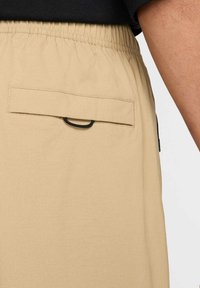 Shorts beiges avec une taille élastique, dotés d'une poche arrière et d'un détail en boucle noire. Le tissu semble lisse et léger.