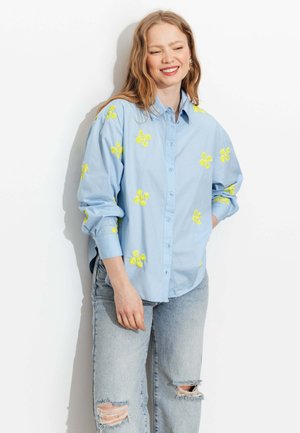 Femme souriante aux longs cheveux portant une chemise bleu clair à boutons avec une broderie de fleur jaune et un jean clair déchiré, debout contre un mur blanc.
