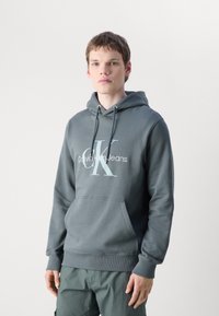 Siva kapuca iz mešavine bombaža, z vtičnim žepom, kapuco s vrvico ter velikim belim napisom "Calvin Klein Jeans".