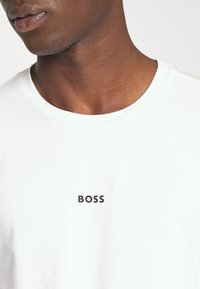 Hvid bomulds-T-shirt med sort "BOSS" logo på forsiden. Rund halsudskæring, korte ærmer og glat tekstur.