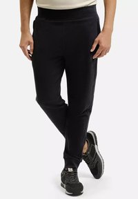 Mężczyzna ubrany w czarne dopasowane dresy i czarne tenisówki, stoi z jedną nogą skrzyżowaną na drugiej na białym tle.