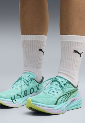 Jambes portant des chaussettes blanches Puma et des chaussures de course Puma bleu clair avec le texte "HYROX" et des semelles jaune-vert sur fond gris.