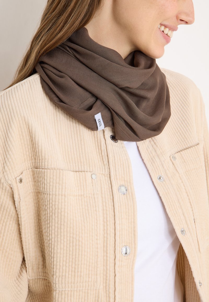 Écharpe légère marron enroulée autour du cou, fabriquée en tissu doux. Portée avec une veste en velours côtelé beige comportant des détails de poches et des boutons métalliques.