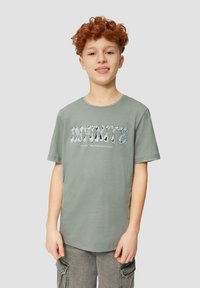 Lichtgroene katoenen t-shirt met de tekst "INFINITE" in golvende blauwe letters, ronde hals, korte mouwen en een ontspannen pasvorm. Grijze cargoshorts.